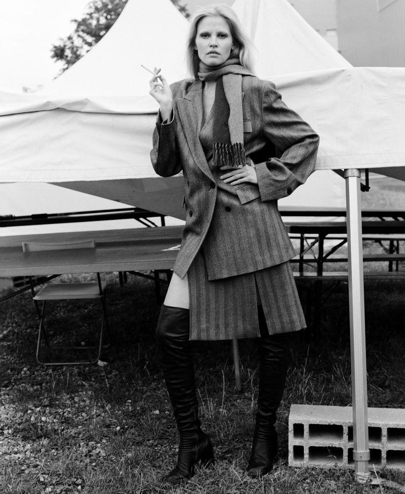 A monochrome fashion shot of an adult female model in a blazer and boots, holding a cigarette and posing outdoors near a tent.