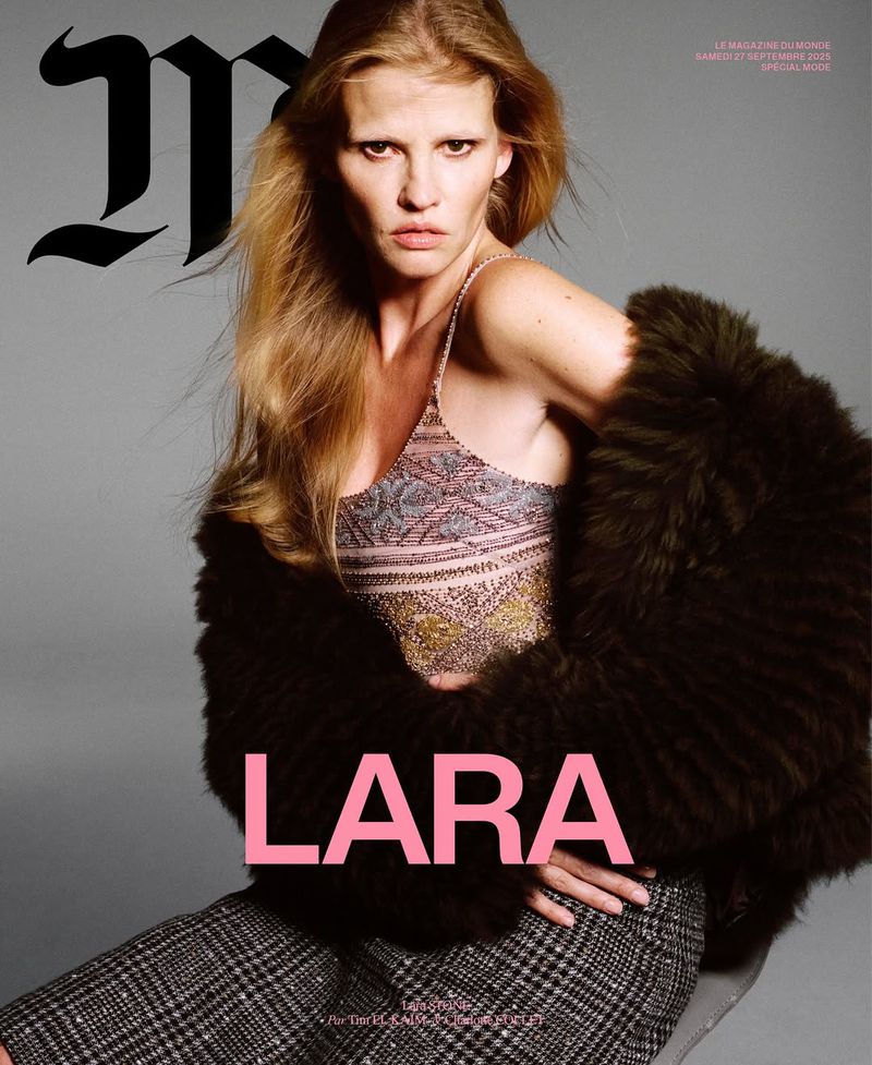 Lara Stone poses for a magazine cover, wearing a tank top, skirt, and fur coat, photographed against a grey studio backdrop.
