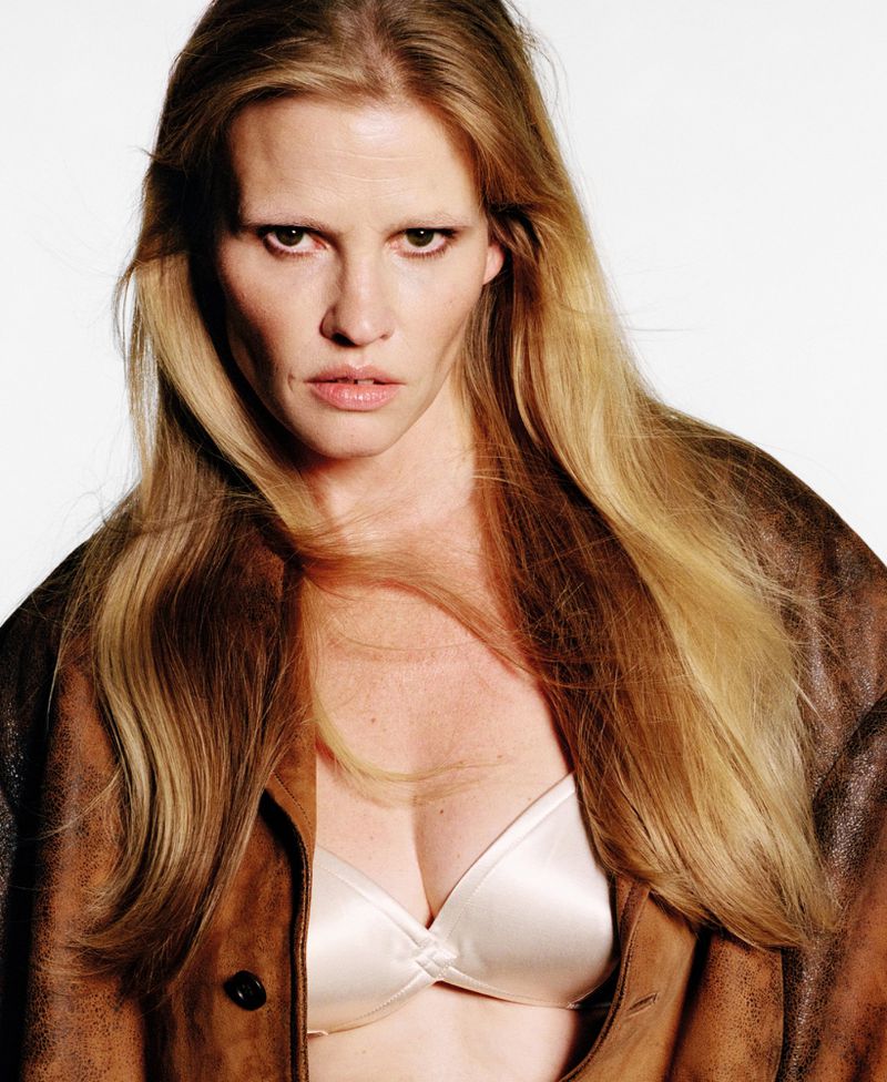 Portrait of an adult female with long blonde hair, wearing a brown leather jacket over a white bra, against a bright white background.
