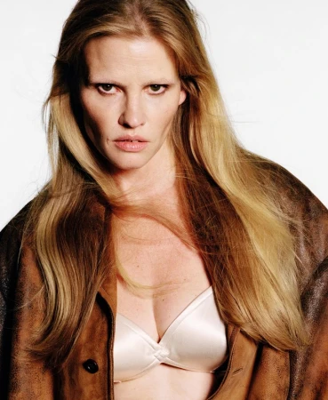 Portrait of an adult female with long blonde hair, wearing a brown leather jacket over a white bra, against a bright white background.