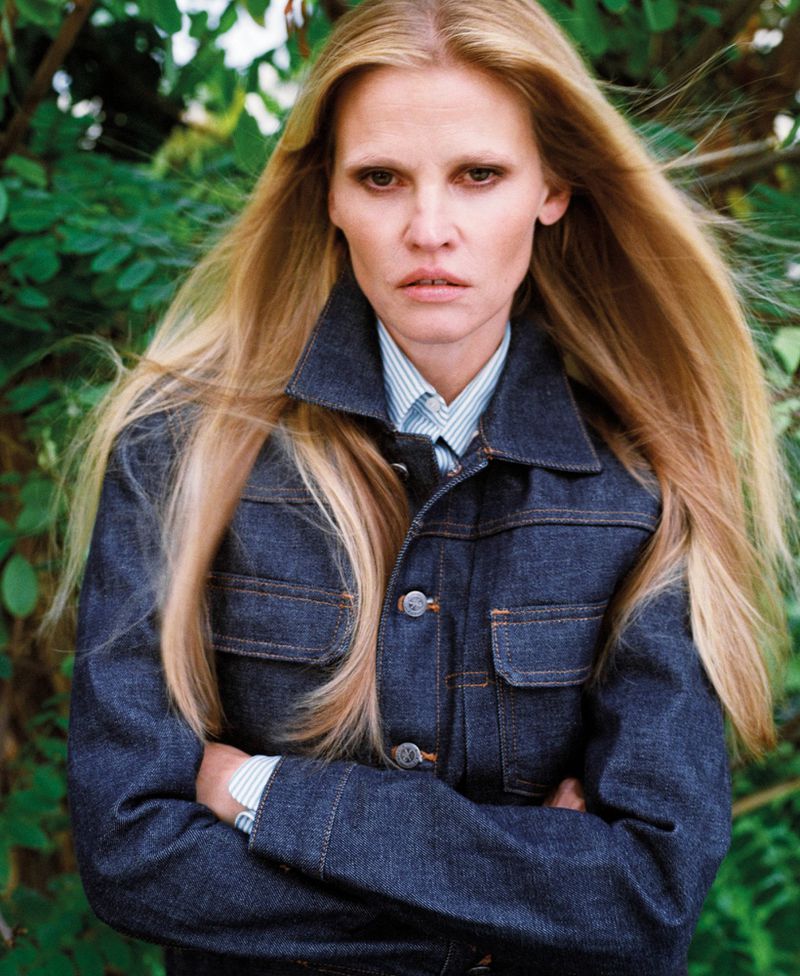 Blonde haired woman wearing a denim jacket and blue striped shirt in front of green foliage. Medium shot of a fashion portrait.