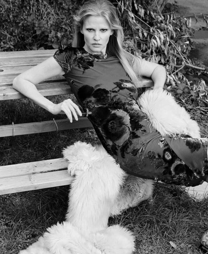 Black and white fashion portrait of a blonde woman reclining on a wooden bench in a patterned dress with a shaggy fur garment.