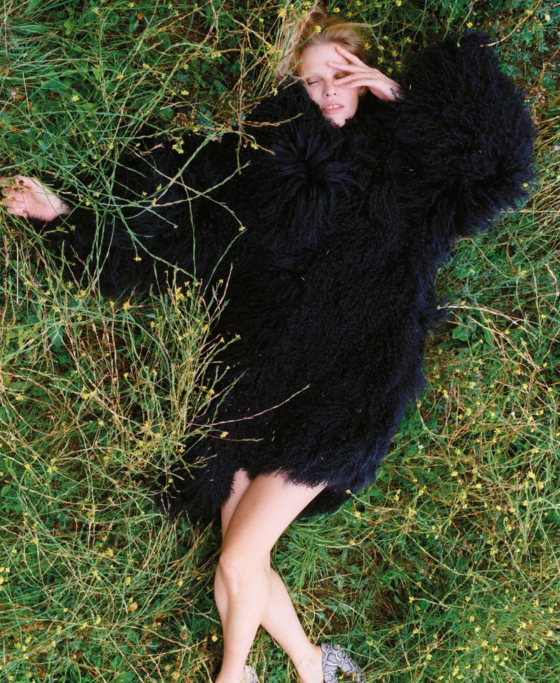 Woman in shaggy black coat rests in grassy field, eyes closed, hand near face, blonde hair spread out, outdoor fashion, daytime.