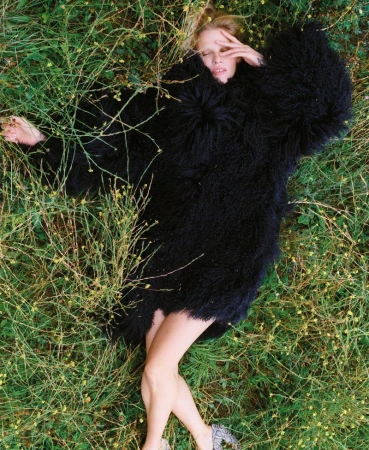 Woman in shaggy black coat rests in grassy field, eyes closed, hand near face, blonde hair spread out, outdoor fashion, daytime.