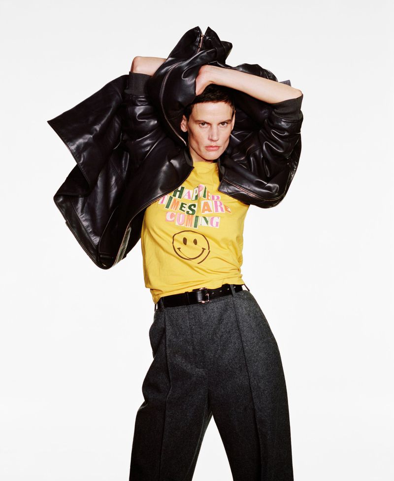 A fashion shot of a young adult androgynous person wearing a yellow shirt with a smiley face and a black leather jacket over their shoulders.