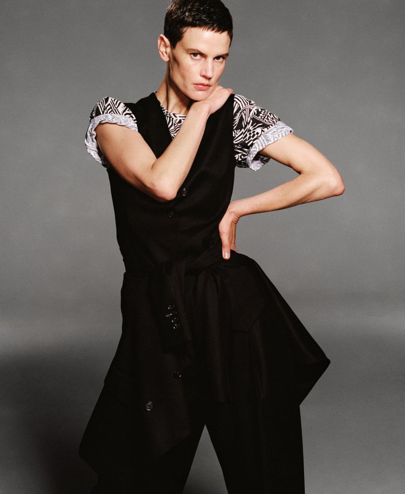 Adult woman with short hair poses in black vest over patterned top and black pants against a neutral studio background in medium shot.