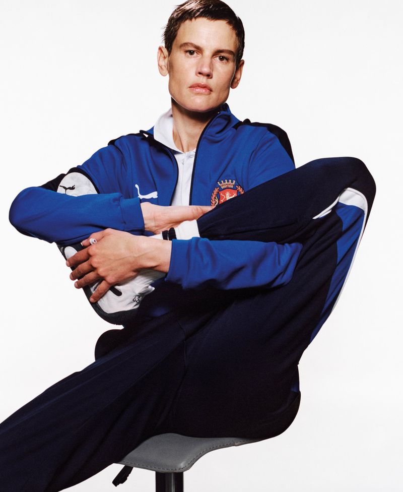 A person sits on a stool with one leg raised, wearing a blue Puma tracksuit against a white background in a studio shot.