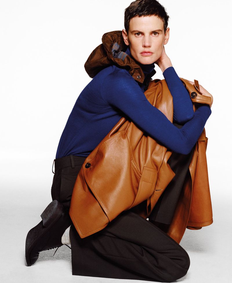 Fashionable person poses in a photo studio wearing a blue sweater, dark trousers, brown leather jacket and shoes against a white backdrop.