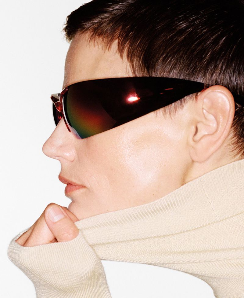 Profile shot of a woman with short dark hair wearing oversized futuristic sunglasses and a beige turtleneck against a white background.