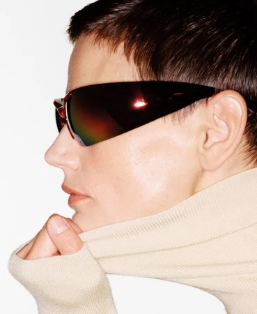 Profile shot of a woman with short dark hair wearing oversized futuristic sunglasses and a beige turtleneck against a white background.