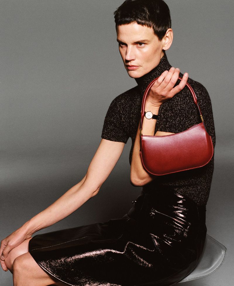 Woman in a black knit turtleneck and shiny skirt sits holding a red-brown handbag in a studio with neutral gray background.