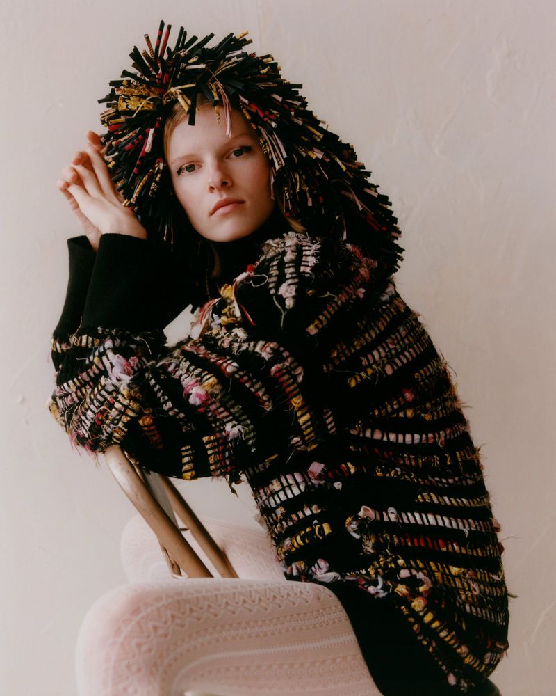 Fashion editorial shot of a blonde female adult wearing a black turtleneck, textured jacket, and headdress while sitting on a wooden chair.