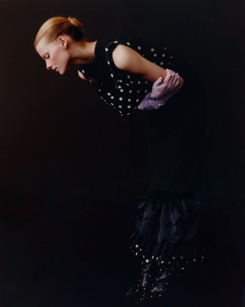 Fashion shot of a woman in a black polka dot dress and lavender gloves against a dark backdrop, looking down with a neutral expression.