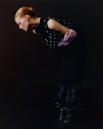 Fashion shot of a woman in a black polka dot dress and lavender gloves against a dark backdrop, looking down with a neutral expression.