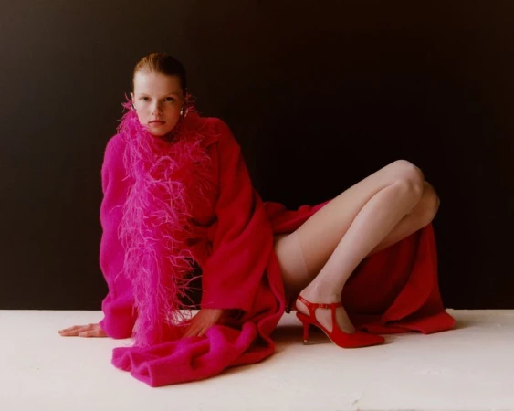 A woman in a hot pink coat with feather trim and red heels poses on a white surface against a dark backdrop in a studio setting.
