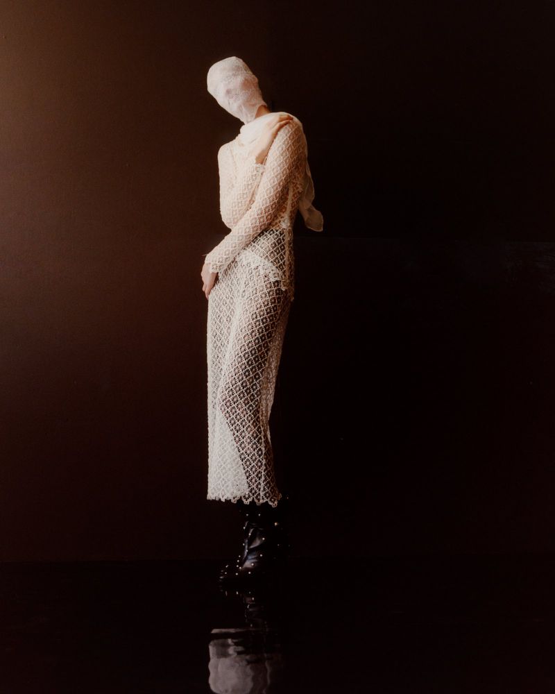 Fashion photo: standing person draped in cream fabric, head wrapped, against a dark brown background with reflective, dark brown floor.