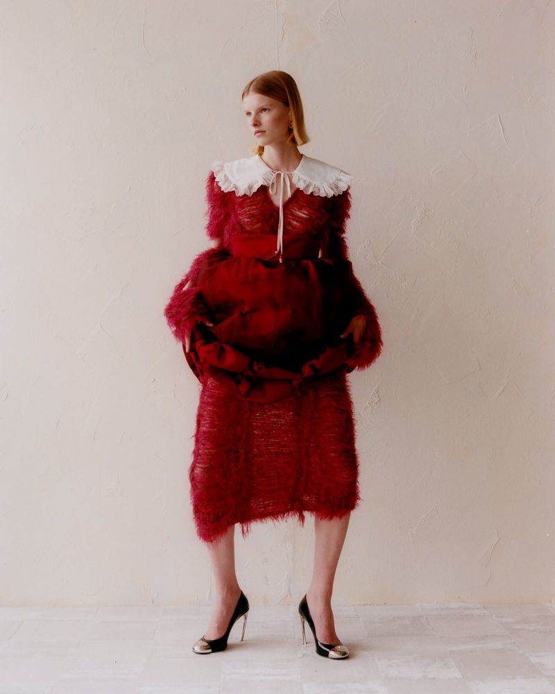 Woman in red textured dress and black heels poses against a neutral wall. The fashion shot features a white collar and unique details.