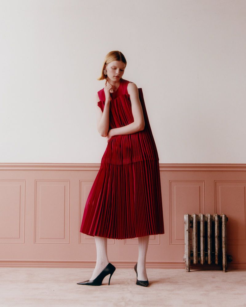 Fashion shot of a slender, light-skinned woman posing in a long red pleated dress, black heels, and sheer white stockings against a pink wall.