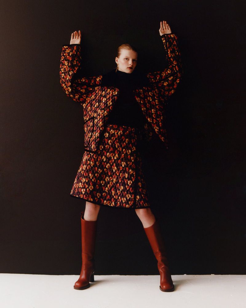 A tall, slender woman with reddish hair, wearing a patterned skirt and jacket, poses with hands up against a dark gray backdrop.