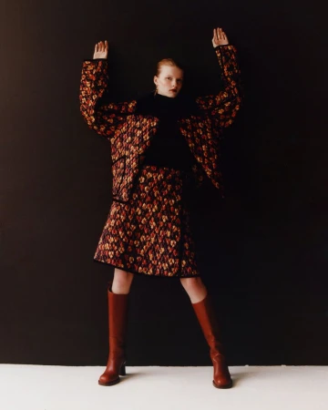 A tall, slender woman with reddish hair, wearing a patterned skirt and jacket, poses with hands up against a dark gray backdrop.