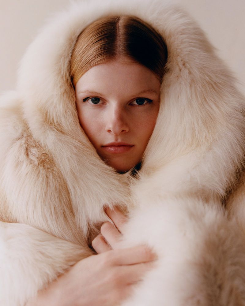 Close-up portrait of a fair-skinned woman with straight red hair wearing a hooded, cream-colored fur coat, against a neutral background.