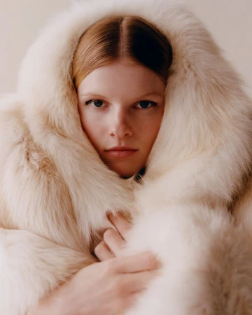 Close-up portrait of a fair-skinned woman with straight red hair wearing a hooded, cream-colored fur coat, against a neutral background.