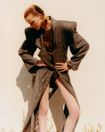Fashion editorial shot of a young adult woman with slicked-back blonde hair wearing a long grey coat with large shoulder pads.