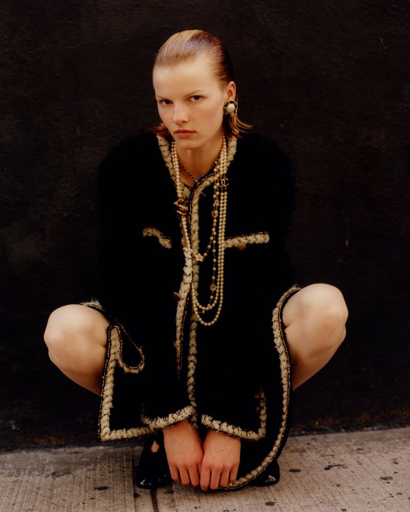 Woman in a black and gold jacket and multiple strands of pearls poses in a crouching position against a dark, textured background.