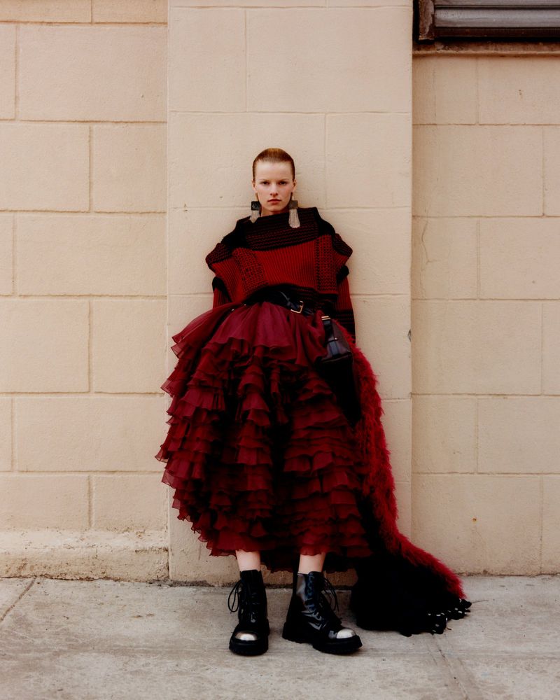 A young adult woman with short red hair poses in a puffy red dress and black boots against a light brick wall in an outdoor setting.