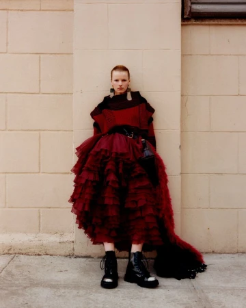 A young adult woman with short red hair poses in a puffy red dress and black boots against a light brick wall in an outdoor setting.