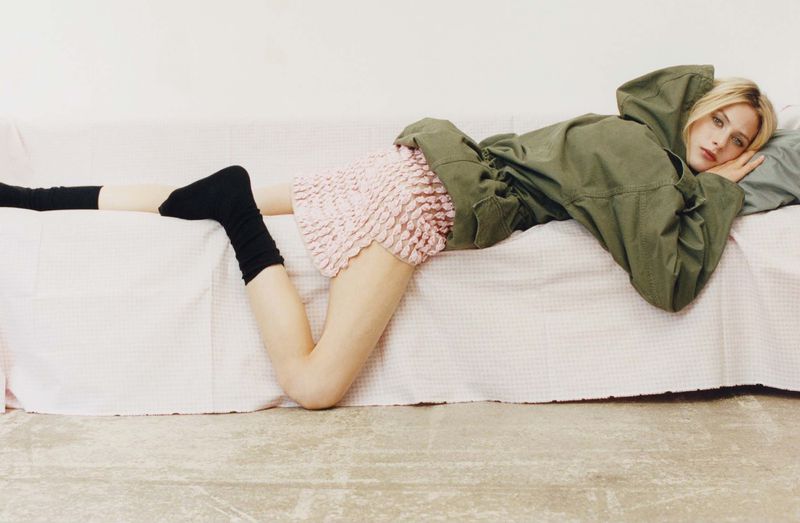 Blonde model in olive hooded jacket and pink ruffled skirt, lying on white couch. She is wearing black socks. Indoor, studio setting.
