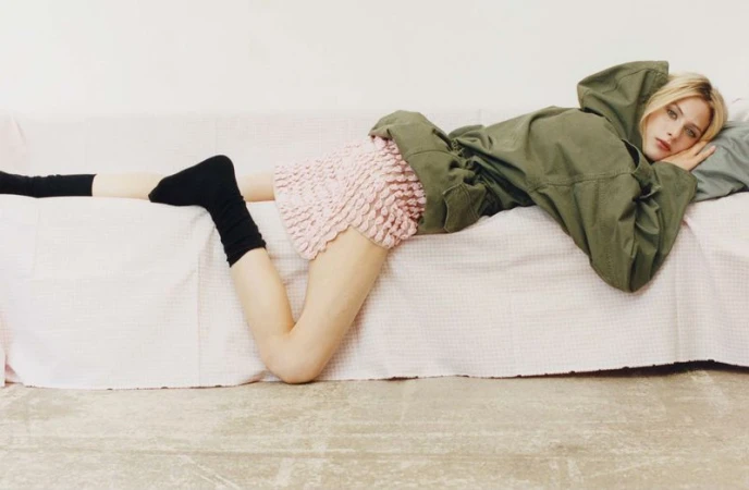 Blonde model in olive hooded jacket and pink ruffled skirt, lying on white couch. She is wearing black socks. Indoor, studio setting.