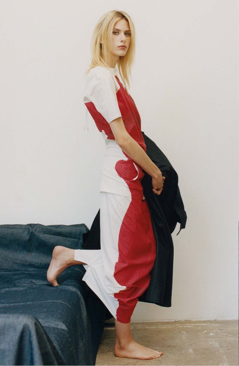 A woman models a white and red top and pants with bare feet, leaning on a covered platform against a white wall in a fashion editorial.