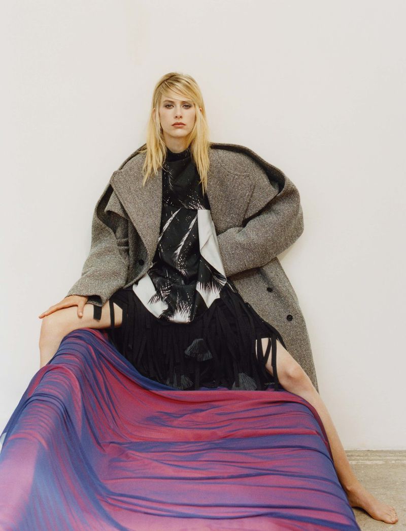 Fashionable blonde woman in grey wool coat poses sitting on draped fabric in an indoor setting against a white wall. Studio lighting.