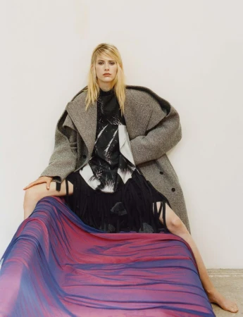 Fashionable blonde woman in grey wool coat poses sitting on draped fabric in an indoor setting against a white wall. Studio lighting.