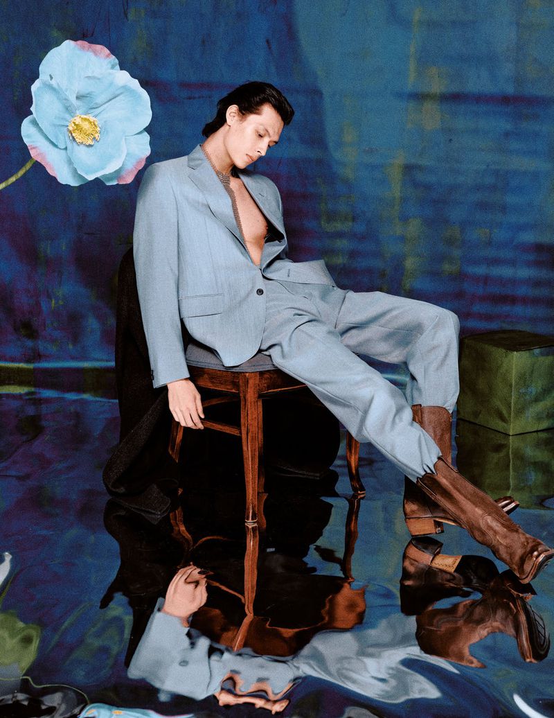 Young adult in a light blue suit sits on a wooden stool in an abstract studio setting with a flower and painterly background.