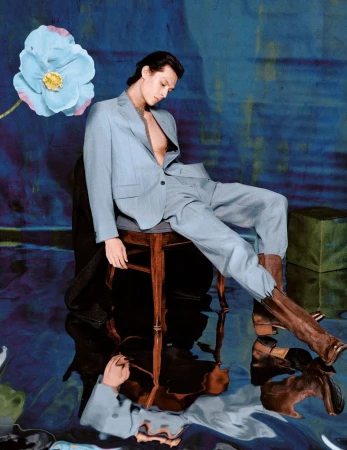 Young adult in a light blue suit sits on a wooden stool in an abstract studio setting with a flower and painterly background.