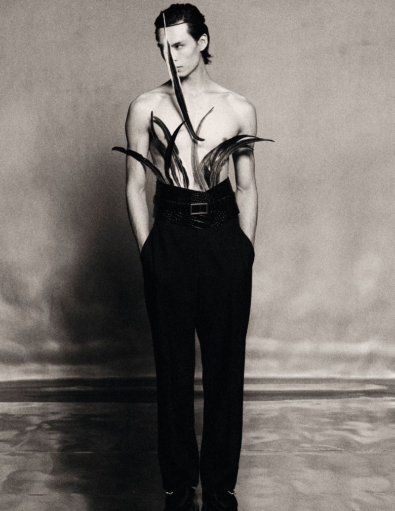 Male fashion model in dramatic studio shot wearing black trousers and a belt, adorned with black feathers around his waist.