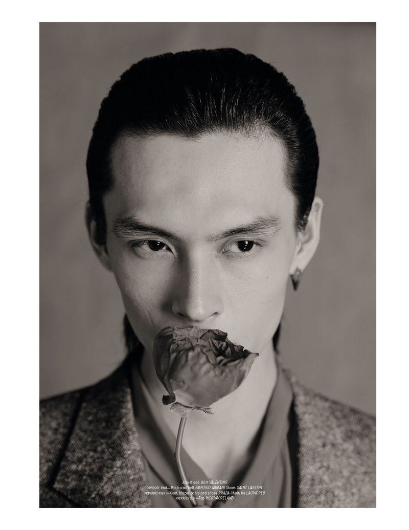 Portrait of a young adult with dark hair holding a flower in their mouth against a neutral background, wearing a blazer and collared shirt.