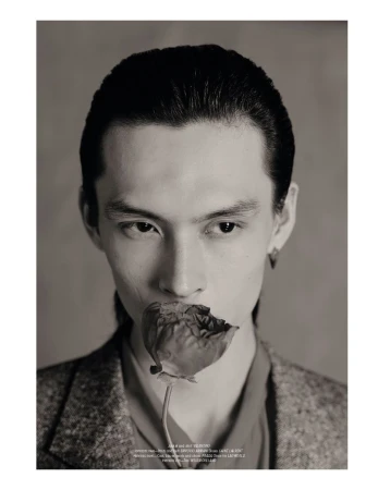Portrait of a young adult with dark hair holding a flower in their mouth against a neutral background, wearing a blazer and collared shirt.