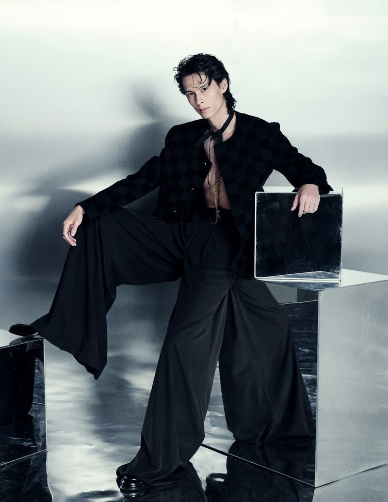A young adult poses in an indoor studio with reflective geometric cubes, wearing an unbuttoned black jacket, wide-legged pants, and jewelry.