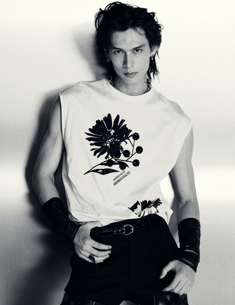 Monochrome studio portrait of a young adult male model with long hair wearing a graphic sleeveless t-shirt, wristbands, and belt.