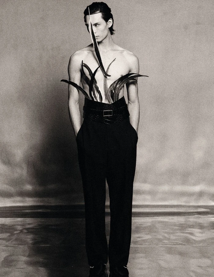 Shirtless male model wearing high-waisted black trousers and sculptural dark feathers tucked into the waistband against a studio backdrop.