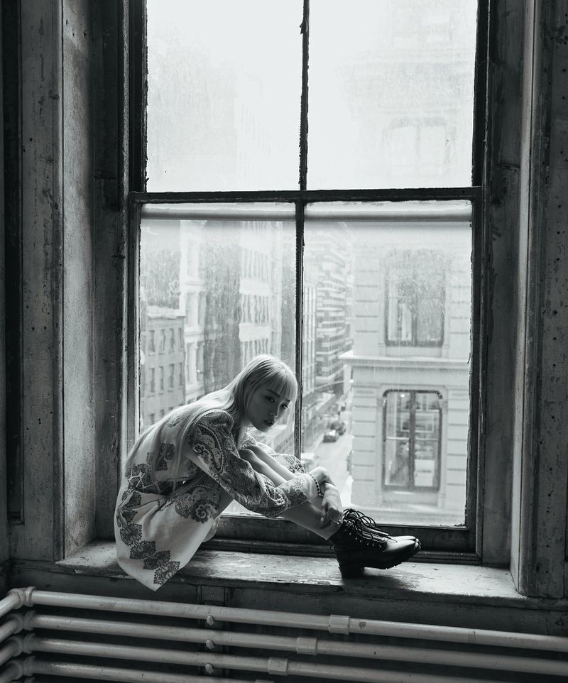 Black-and-white portrait of a woman with short blonde hair, sitting by a window overlooking a city, wearing a patterned outfit.