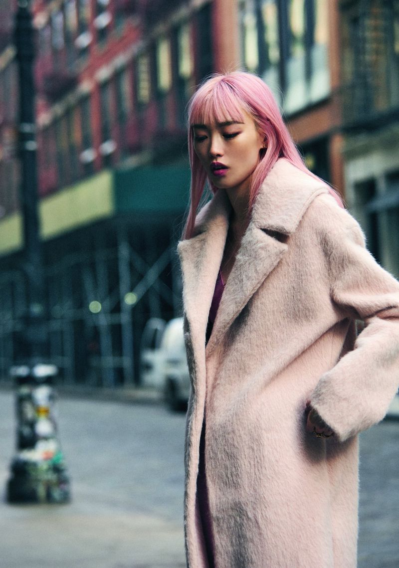 A woman with pink hair walks down a city street, wearing a pink coat, with buildings and a parked car visible in the background.