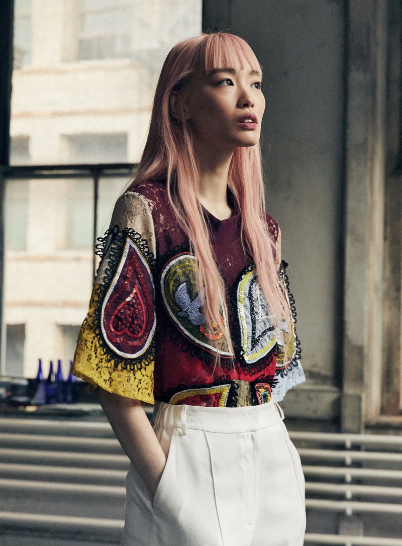 Young woman with pink hair in printed top and white pants poses indoors with hands in her pockets near windows.