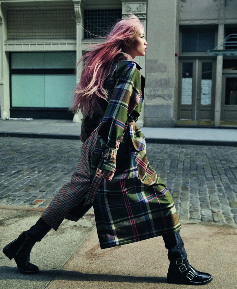 Woman with pink hair in a plaid trench coat walks on a city street with architectural details. Street-style fashion editorial.