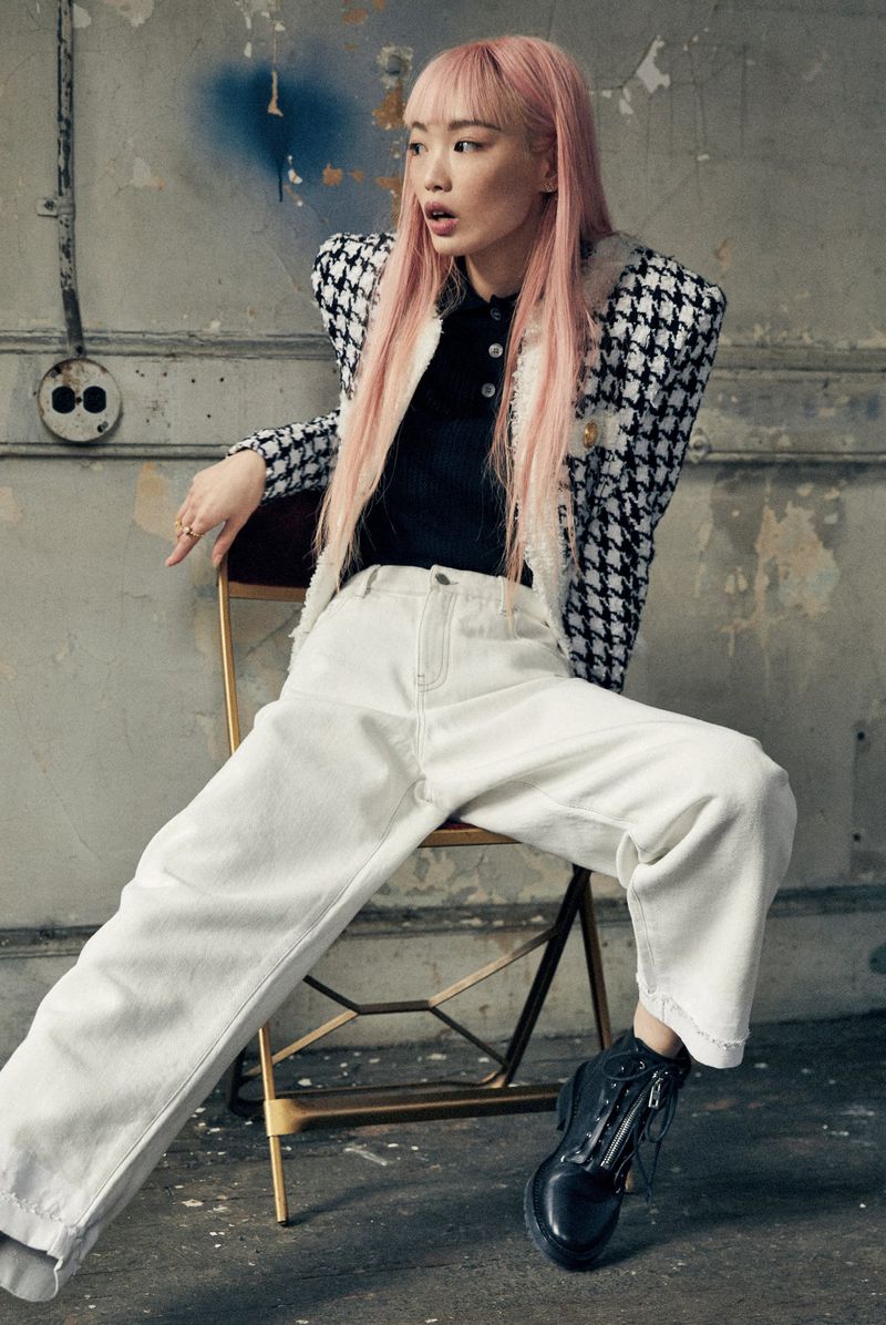 Model Fernanda Ly with pink hair wearing houndstooth jacket and white trousers seated on chair against distressed wall