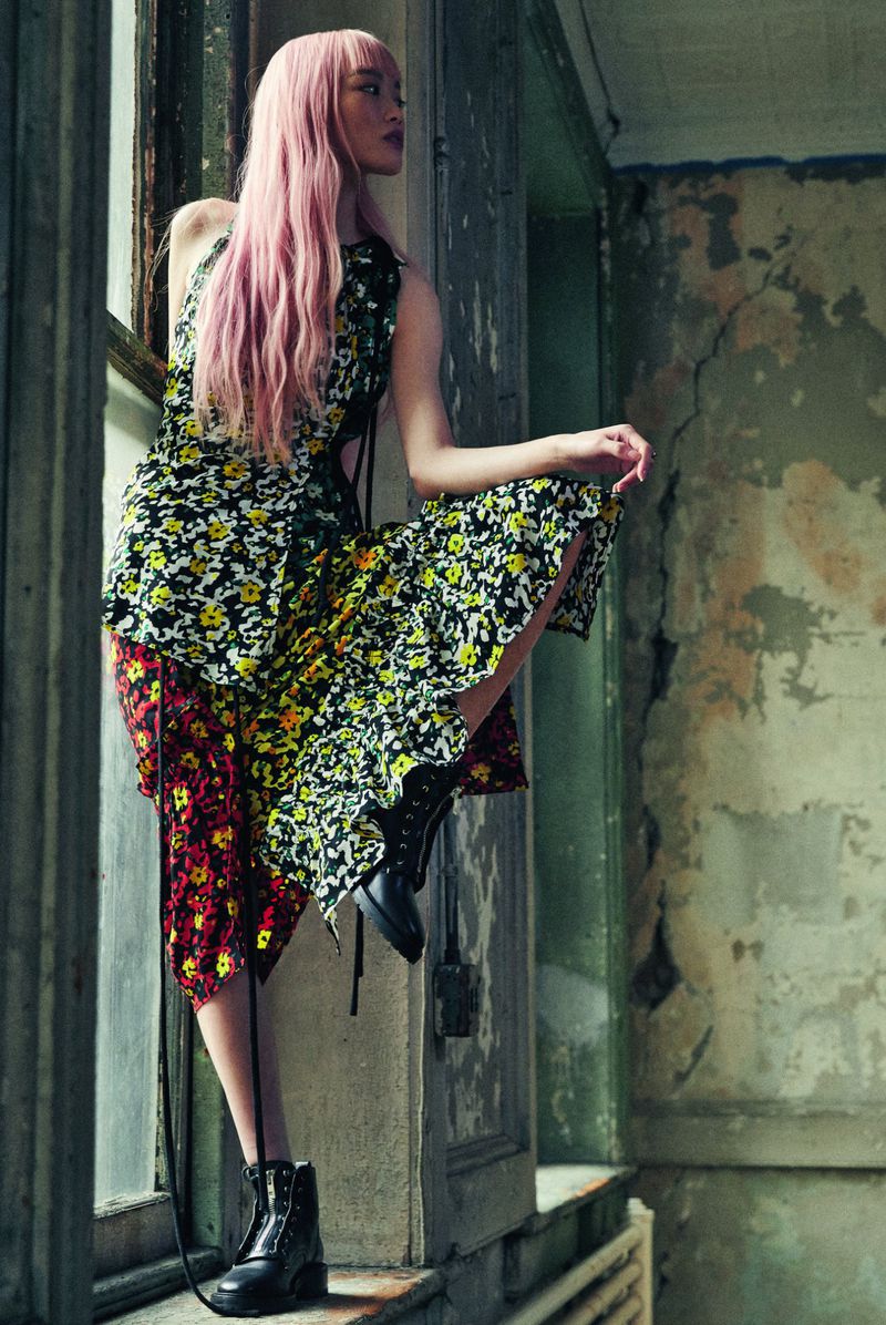 Young woman with pink hair poses near a weathered window in a floral print dress and combat boots in a fashion editorial setting.