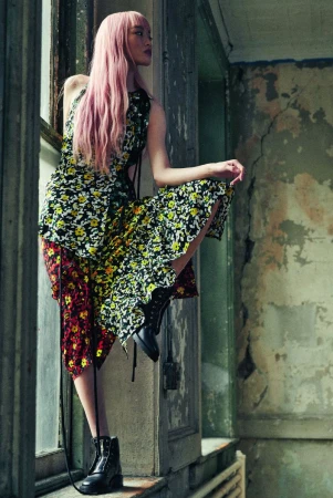 Young woman with pink hair poses near a weathered window in a floral print dress and combat boots in a fashion editorial setting.